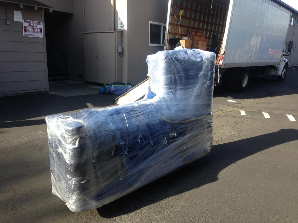 Couch fully wrapped in moving blankets and stretch wrap, waiting on the pavement beside an open moving truck
